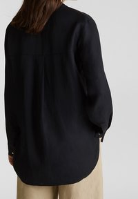 Person mit langen braunen Haaren, die ein lockeres schwarzes Langarmshirt und beiges Hosen trägt, von hinten vor einem einfarbigen Hintergrund gezeigt.