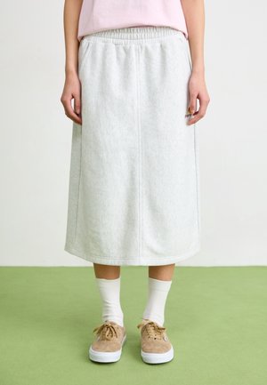 Persoon draagt een lichtgrijze knielange rok, witte sokken, beige sneakers en een lichtroze top, staand op een groene vloer tegen een witte muur.