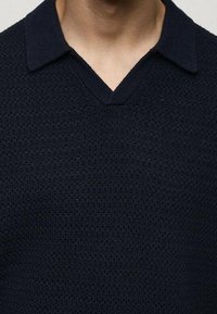 Chemise en maille texturée bleu marine foncé avec un col en V portée par une personne.