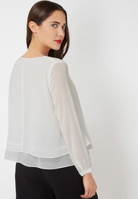 Blusa blanca, transparente, con mangas largas, dobladillo en capas y puño abotonado. Textura suave; corte relajado; diseño minimalista.