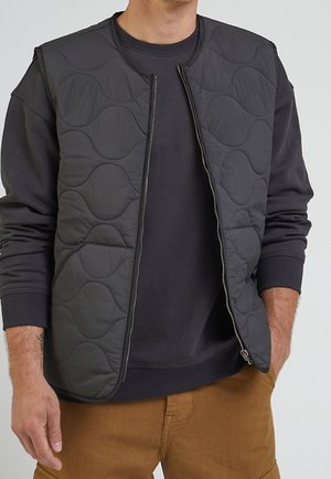 Man draagt een zwart doorgestikt bodywarmer over een zwart sweatshirt en een bruine broek, met één hand in de zak en het bodywarmer deels dichtgezipped.
