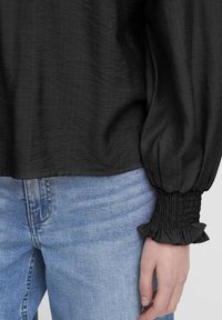 Blusa nera con texture e maniche voluminose, caratterizzata da polsini a sbuffo. Abbinata a jeans di denim azzurro chiaro.
