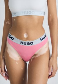 Rosa truse med grå HUGO liv, med marineblå tekst. Glatt stoff, høy-kuttet design, minimal dekning og ingen synlige mønstre.