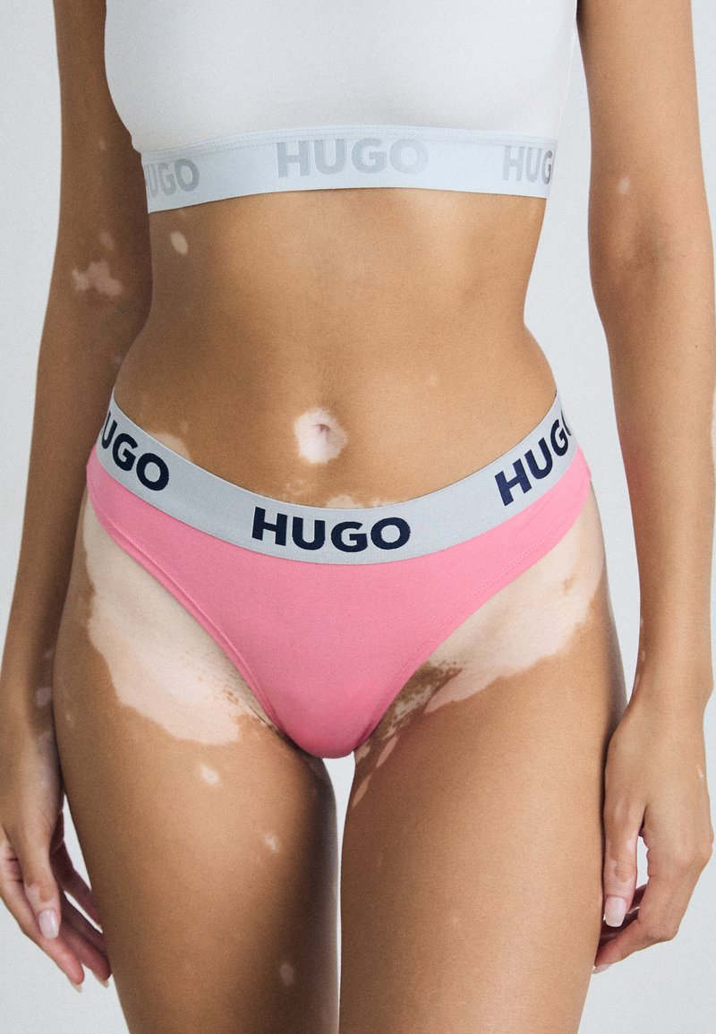 Rosa truse med grå HUGO liv, med marineblå tekst. Glatt stoff, høy-kuttet design, minimal dekning og ingen synlige mønstre.