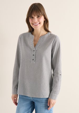Femme souriante portant une chemise à manches longues rayée verticalement noir et blanc et un jean bleu, debout devant un fond beige uni.