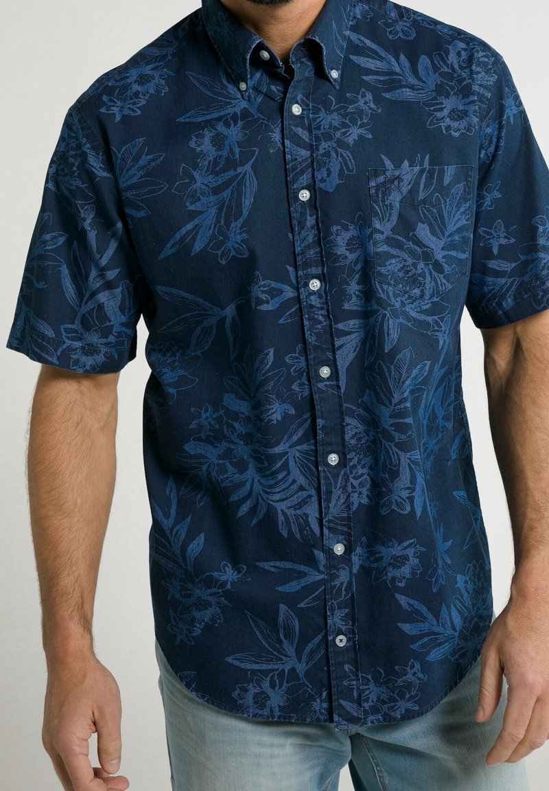 Muž v námořnicky modré košili s krátkým rukávem a jemným květinovým vzorem, v světle modrých džínách.