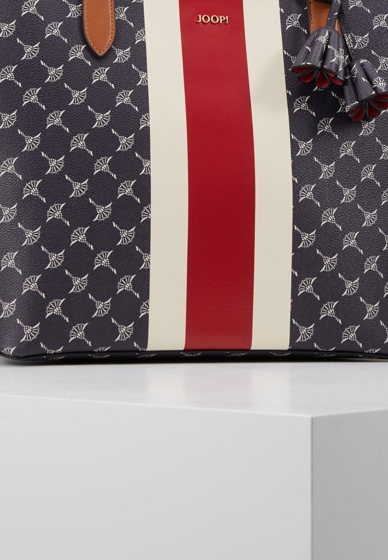 Modrá vzorovaná taška přes rameno s krémovým a červeným středovým pruhem, hnědými koženými rukojeťmi a střapcovými ozdobami. Vyrobená z texturovaného syntetického materiálu.