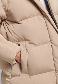 Beige, polstret jakke med en glat, quiltet tekstur, store sidelommer og en høj krave, der giver varme og dækning.