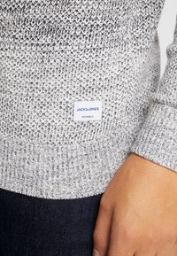 Stickad tröja i grått och vitt. Har ribbade muddar och nederkant. Etikett synlig med "JACK & JONES ORIGINALS"-logotyp. Mjuk textur.