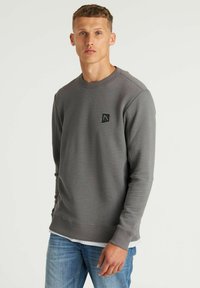 Giovane uomo con capelli biondi corti che indossa una felpa grigia a coste e jeans blu su uno sfondo bianco uniforme.