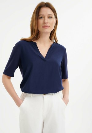 Top bleu marine à manches courtes avec un design en V, fabriqué en tissu lisse. Associé avec un pantalon taille haute blanc comprenant des poches latérales.