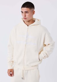 Sudadera cremosa con cremallera de tela suave, que presenta un logo frontal en blanco. Incluye capucha y bolsillos tipo canguro con puños acanalados.