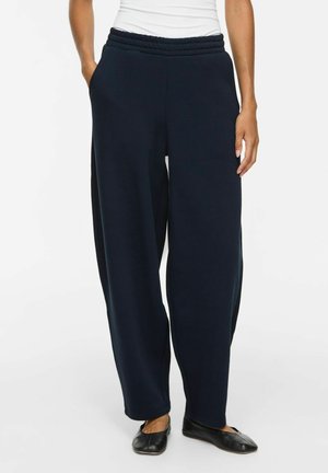 Femme portant un pantalon ample bleu marine foncé avec une taille élastique et des chaussures plates noires sur fond blanc.