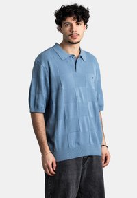 Camisa polo de color azul claro con textura a cuadros, de manga corta y cuello tipo polo. Combinada con unos vaqueros sueltos de color gris oscuro.
