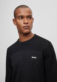 Črna pletena jopica z teksturiranim zgornjim delom in rebrastim ovratnikom, z belim logotipom "BOSS" na prsih.