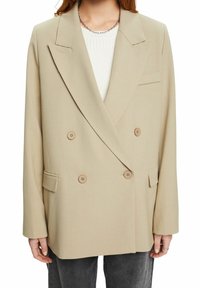 Blazer beige doppiopetto con grandi bottoni, reverse a lancia e tasche laterali. Tessuto morbido e vestibilità rilassata, mostrato sovrapposto a una maglietta bianca.