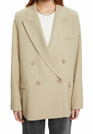 Beiger doppelt-knöpfiger Blazer mit großen Knöpfen, Reverskragen und seitlichen Taschen. Glattes Material und lässige Passform, präsentiert über einem weißen Oberteil.