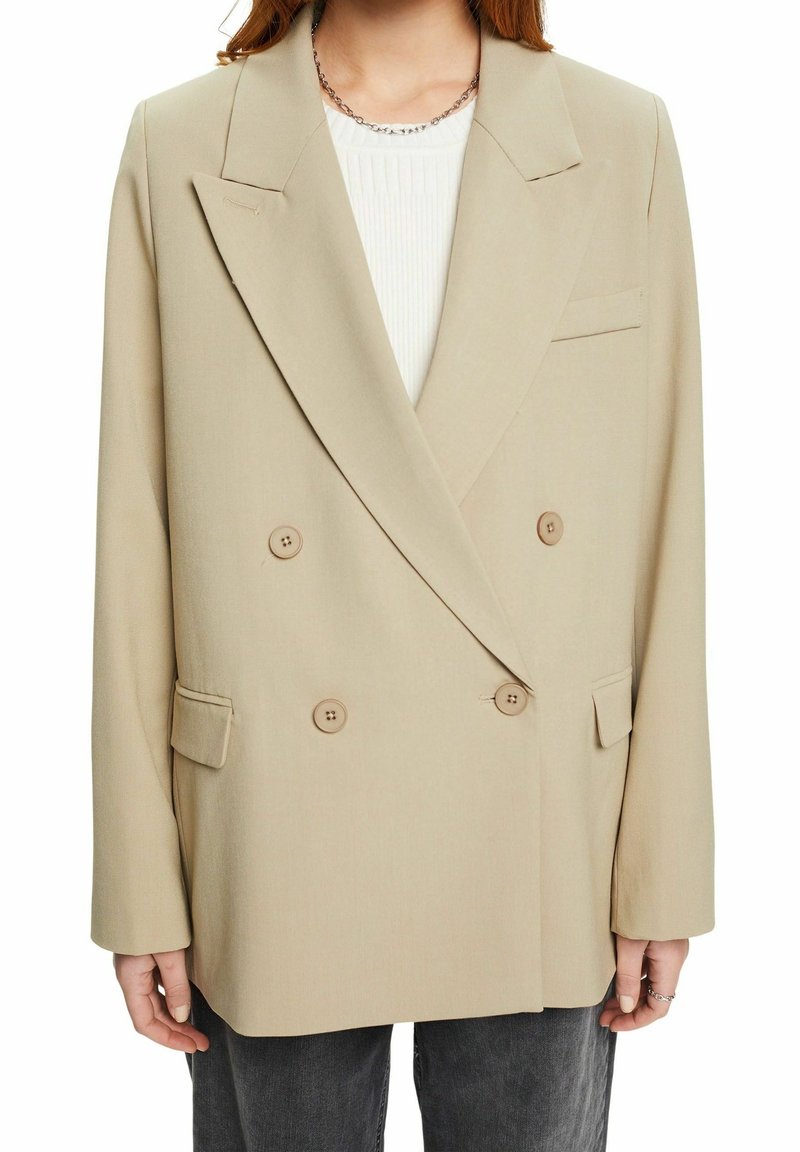 Blazer beige doppiopetto con grandi bottoni, reverse a lancia e tasche laterali. Tessuto morbido e vestibilità rilassata, mostrato sovrapposto a una maglietta bianca.