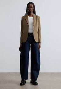 Beige skræddersyet blazer med to knapper, båret over en hvid tætsiddende top og mørkeblå vidde bukser, kombineret med sorte læderankelstøvler.