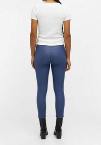 T-shirt blanc ajusté et leggings bleus taille haute, avec une texture lisse. Bottines noires à talon épais.