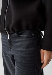 Felpa nera con zip, dalla texture liscia e orlo elastico, abbinata a jeans grigio scuro con un sottile motivo a righe sul tessuto.