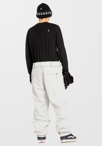 Chemise noire à manches longues, pantalon large blanc, bonnet en tricot noir avec logo, gants noirs, et baskets noires avec accents de logo blancs.