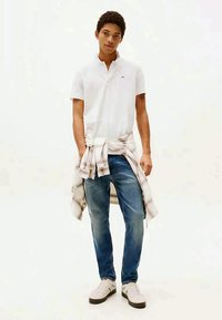 Young man wearing white polo shirt, blue jeans, white sneakers, with plaid shirt tied around his waist, standing against plain background.