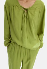 Zielona lniana bluzka z dekoltem w kształcie litery V i detalami w postaci wiązań, z długimi rękawami i marszczonymi mankietami, w komplecie z pasującymi spodniami z elastycznym pasem.
