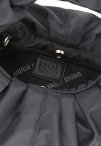 Interiér černé textilní tašky s plisovaným designem, zlatými detaily a koženou nálepkou s embosovaným nápisem "KAZAN STUDIO." Hladká textura.