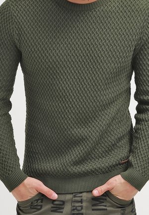 Uomo che indossa un maglione lavorato a maglia verde con le mani nelle tasche dei pantaloni verdi con lettere nere stampate.