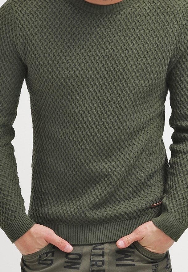 Homme portant un pull en maille verte texturée avec les mains dans les poches d'un pantalon vert avec des lettres noires imprimées.
