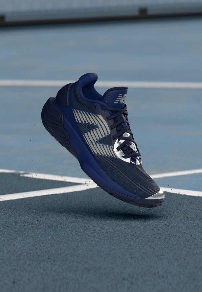 Chaussure de sport navy au design dynamique, tige en maille texturée, accents réfléchissants et semelle rembourrée. Flotte au-dessus d'une surface de court bleue.