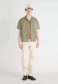 Chemise à manches courtes de couleur vert olive, débardeur marron en dessous, pantalon crème et bottines noires sous la cheville, le tout sur fond uni.