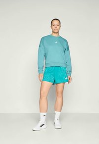 Tirkīzzils kapucis ar baltu Adidas logo, kopā ar tirkīzzilām rakstainām šortiem. Balti sporta zeķes un kedas noslēdz izskatu.