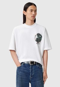 Tricou alb din bumbac cu mânecă scurtă, cu un grafic de șarpe verde și accente florale pe față. Asortat cu blugi albaștri și o curea neagră.