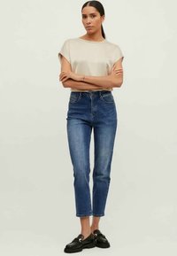 Nő áll karba tett kézzel, bézs rövid ujjú felsőt, magas derekú kék farmert és fekete mokaszint visel, egyszerű háttér előtt.