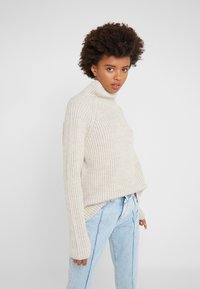 Beige ribbed turtleneck sweater with long sleeves, soft texture, slightly loose fit, paired with light blue jeans, standing against a plain backdrop.