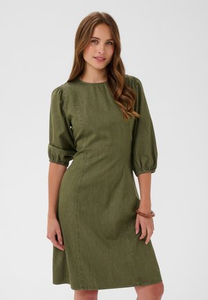 Femme aux longs cheveux bruns portant une robe olive, longueur genoux, à manches bouffantes jusqu’aux coudes, debout devant un fond clair uni.