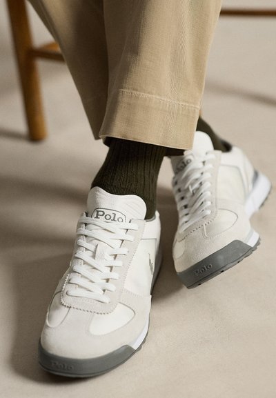 Białe i beżowe sneakersy Polo z szarymi podeszwami, noszone z ciemnozielonymi skarpetkami i beżowymi spodniami, osoba siedząca ze skrzyżowanymi nogami.