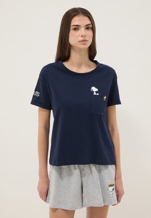Giovane donna che indossa una maglietta blu navy con grafiche di Snoopy e Woodstock e pantaloncini grigi con Snoopy che porta Woodstock in un nido.