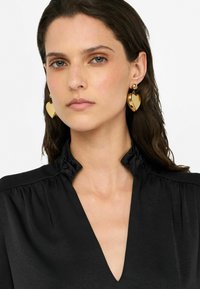 Orecchini a forma di cuore in oro con finitura lucida, attaccati a una piccola palla d'oro, indossati con una blusa nera con colletto a balze.
