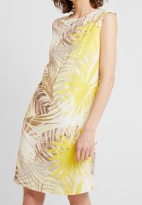Frau in einem ärmellosen Kleid mit gelbem und braunem tropischen Blattmuster und kleinen dekorativen Nieten am Ausschnitt vor weißem Hintergrund.