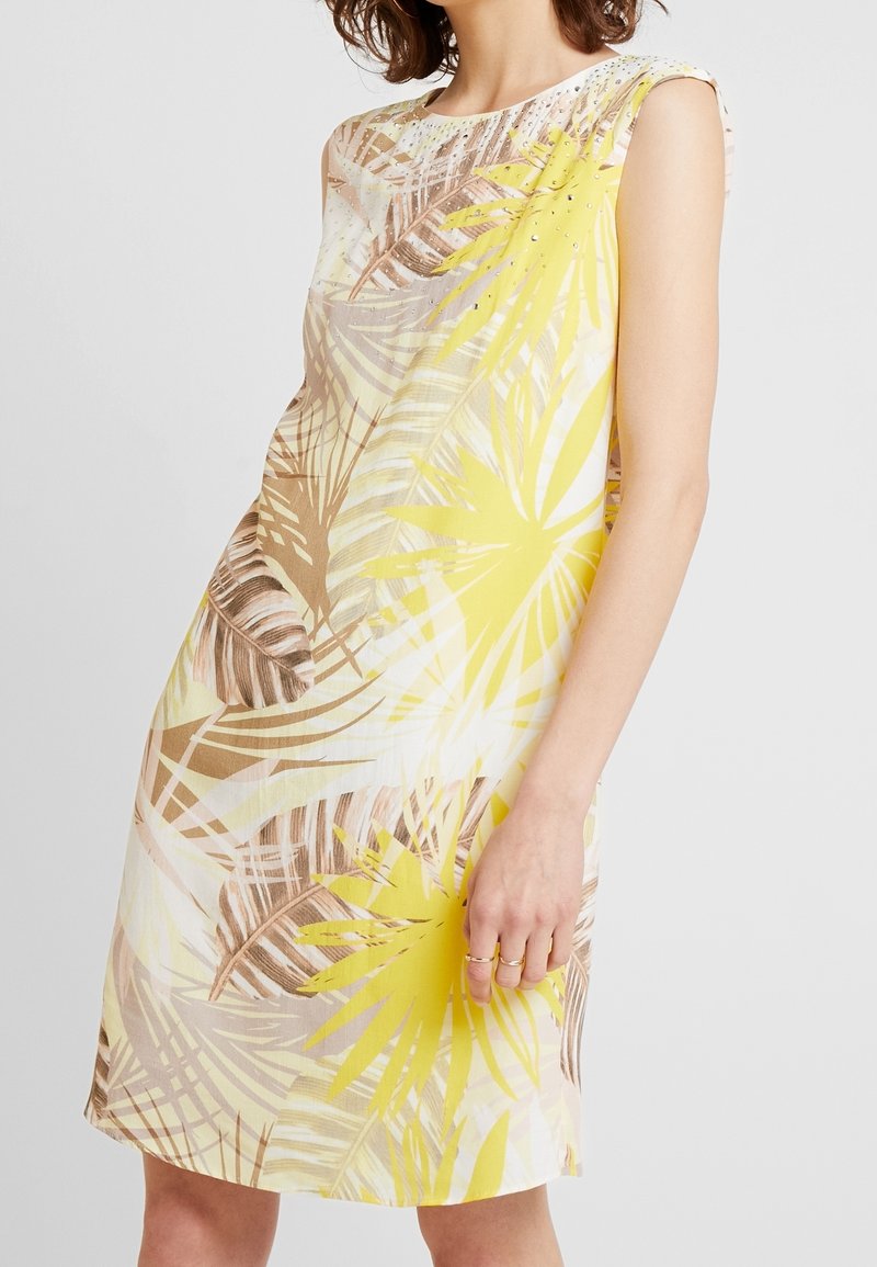 Frau in einem ärmellosen Kleid mit gelbem und braunem tropischen Blattmuster und kleinen dekorativen Nieten am Ausschnitt vor weißem Hintergrund.