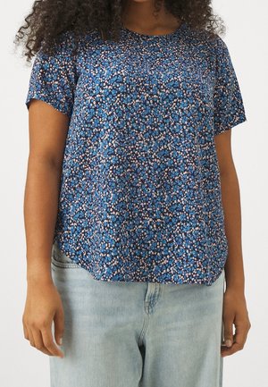 Mujer con blusa azul de manga corta con estampado floral combinada con vaqueros azul claro, de pie frente a un fondo liso.