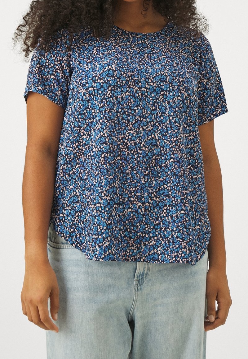 Femme portant un chemisier bleu à manches courtes avec un imprimé floral, associé à un jean bleu clair, debout devant un fond uni.
