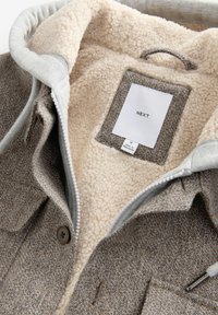 Ruskosa herringbone-takki, jossa on kerma fleece-vuori, harmaa huppu, ruskeat nappiat ja sisällä ommeltu etiketti. Vetoketju näkyvissä.