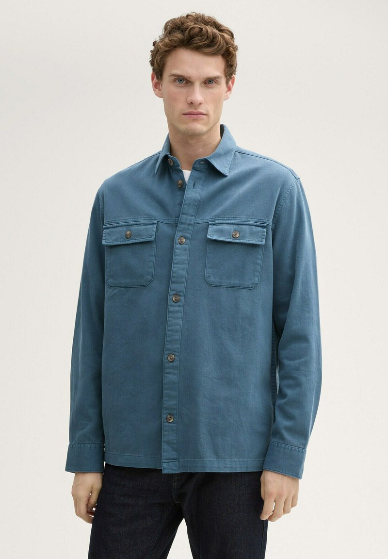 Camicia blu con bottoni in morbido tessuto di cotone; presenta due tasche sul petto, colletto corto e un orlo dritto. Indossata con jeans scuri.