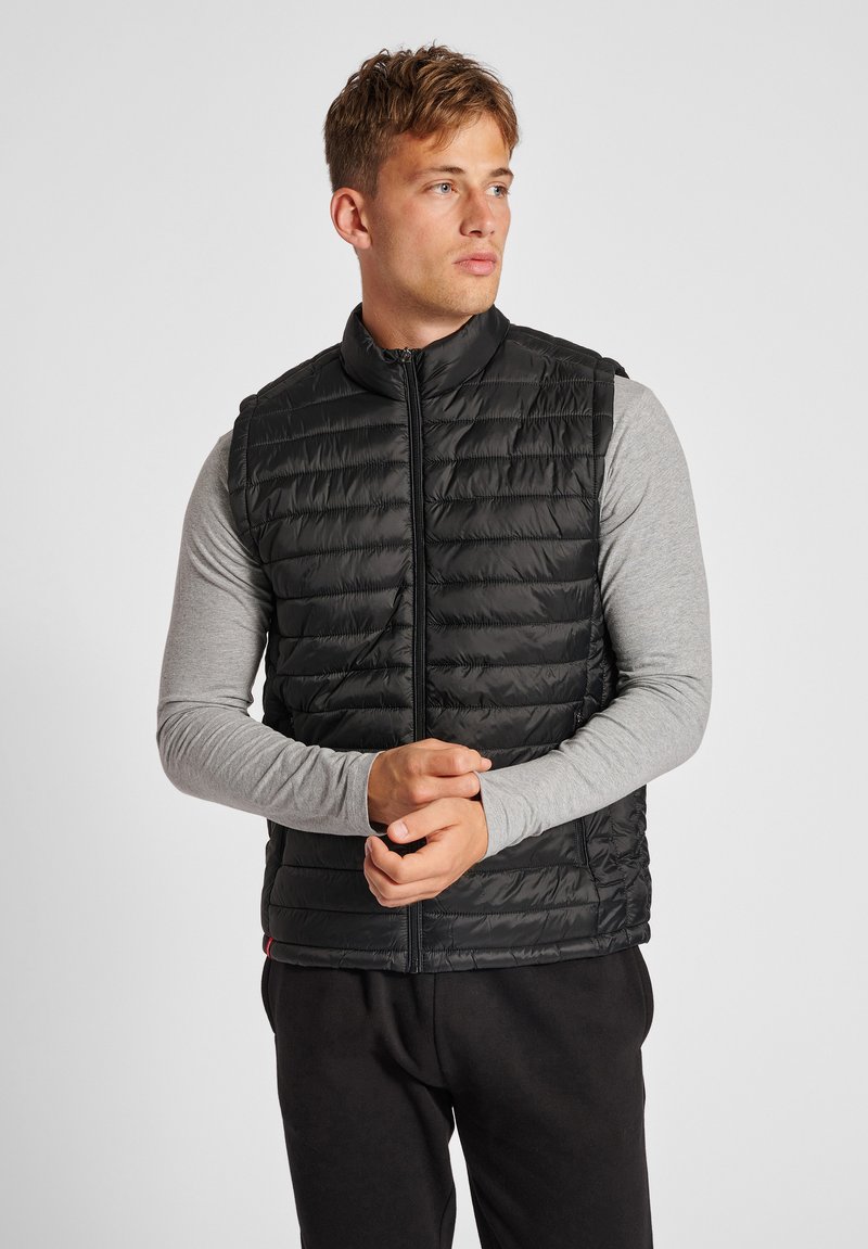 Mann trägt schwarze gesteppte Weste über hellgrauem Langarmshirt, steht mit verschränkten Händen und blickt nach rechts vor einfachem Hintergrund.