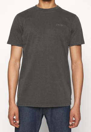 Homme portant un t-shirt noir délavé à col rond avec un petit logo "Han Kjøbenhavn" sur le côté gauche de la poitrine et un jean bleu foncé.