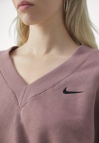Růžová mikina s výstřihem do V vyrobená z měkké látky s žebrovaným límcem a malým černým logem Nike na levé straně. Hladká textura.